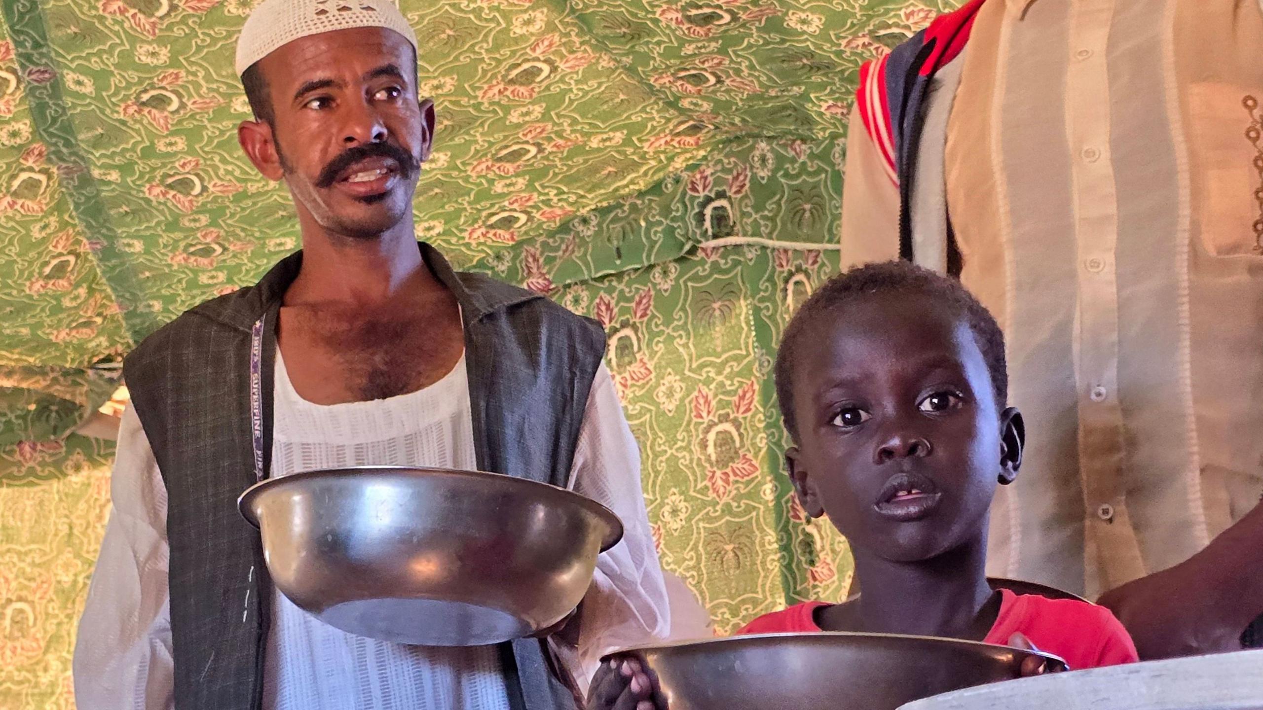 A man in a white robe, white knitted hat and grey coat holds a bowl as he waits for aid alongside a young boy in a red T‑shirt